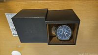 Full view of the Michael Kors Gage Chronograph watch in its box showing the gunmetal-tone bracelet and blue dial with chronograph features.