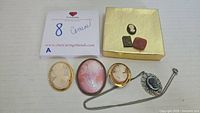 All items laid out: two gold-tone brooches, one large unframed cameo, pendant on chain, loose stones, note card