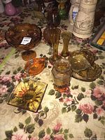 Overview photo of multiple amber-colored vintage art glass pieces including bowls, vases, dishes, and compote on floral fabric background.