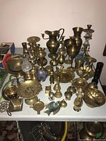 Wide view showing a variety of brass items including vases, bowls, goblets, candle holders, bells, and a pitcher on a table.
