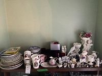 Wide view of stacked china plates, ruby glassware, assorted teacups, porcelain and ceramic figurines, and miniatures arranged on a wooden shelf.