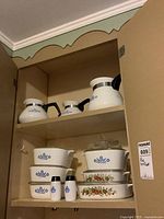 Photo of kitchen cupboard shelf displaying three Corning coffee pots with black handles on upper shelf; lower shelf contains Blue Cornflower and Spice of Life Corning casseroles with lids, and Blue Cornflower salt and pepper shakers.
