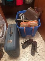 Blue plastic bin containing various vintage handbags and clutches including woven wicker handbag, black and white woven handbag, dark brown textured handbag, and black leather clutch. Next to bin is vintage dark blue hard case suitcase and pair of brown ankle boots size 7 on floor.
