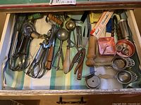 Drawer of assorted vintage kitchen utensils including egg beaters, whisk, ice cream scoops, nut cracker, food decorator box, tea strainers, small molds.