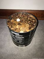 Metal container filled with mixed USA and Canadian pennies placed on a carpeted floor near wooden baseboard.