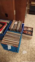 Three boxes: two blue crates with vinyl records and one white box with CDs on a tiled floor near wooden paneling.