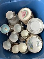 Top-down view of mixed unpaired teacups and stacked saucers with floral motifs and gold trim inside blue container.