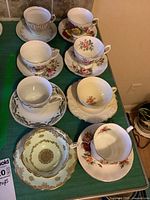 Eight sets of fine English china teacups and matching saucers arranged on a green cloth surface showing varying floral and gold trim patterns.