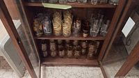 Two shelves of assorted vintage drinking glasses inside wood and glass cabinet. Top shelf holds clear glasses with floral and gold designs; middle shelf holds amber-tinted glasses with gold stripes; bottom shelf has mixed clear and amber glasses.