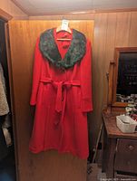Vintage red wool coat with black mink collar hanging on a wooden door. Coat has a matching waist belt and button closure.