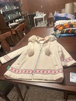 Beige wool coat with fur collar and pom-poms, embroidered sleeve and hem pattern, displayed flat on table