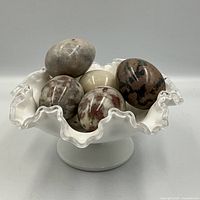 Five polished stone eggs displayed inside a white milk glass footed candy dish with a ruffled and clear-edged top.