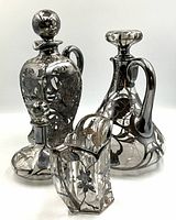 Group view of three Art Nouveau glass items with ornate silver overlay, including creamer and two bottles with stoppers