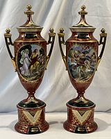 Front view of both porcelain lidded urns side by side showing painted cartouches, handles, lids and pedestal bases