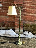 Full view of the vintage brass floor lamp with a bridge arm and cream fabric shade, standing upright outside on pavement against a brick wall.