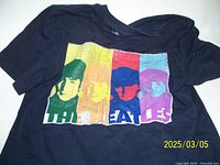 Black T-shirt laid flat showing colorful pop-art portraits of The Beatles across the chest with text below.