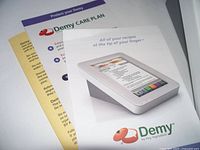 The main photo showing the Demy Recipe Tablet propped up with its screen displaying a recipe interface, surrounded by printed care plan brochures for the device.