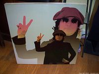 Square board with pop art style John Lennon print showing him with sunglasses, red hat, and peace signs.