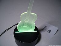 Guitar-shaped LED lamp on black round base lit green with visible embroidered guitar details