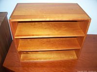 Wooden storage shelf with three slanted shelves inside a rectangular frame, medium-light wood tone and visible wood grain, photographed on a wooden surface.