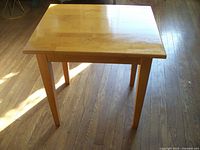 Photo of a wooden square table with a smooth wood finish and natural wood grain. The table stands on four tapered legs on a hardwood floor.