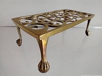 Angled side view of the brass trivet showing ornate scrollwork and claw feet legs