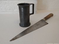 Photo showing antique French pewter measuring tankard with handle alongside long chef's knife with wooden handle and metal blade.