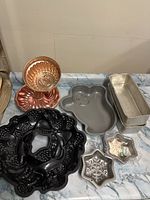 Photo shows various bakeware moulds including copper bundt pans, a black decorative bundt pan, a gray gingerbread man pan, metal loaf pans, and ceramic star dishes on a countertop.