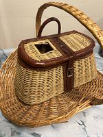 Fishing creel basket with leather trim and latch closure on top, placed on large woven basket.