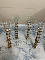 Four glass candlestick holders arranged upright on a marble surface showing their stacked gold rings on clear glass stems
