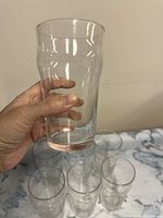Hand holding one clear glass tumbler showing full shape and size with several similar glasses on surface below.