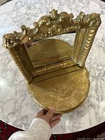 Front view of the gold vanity mirror with the hinged jewelry storage compartment open, showing the mirror and base with gold finish and ornate decorative details.