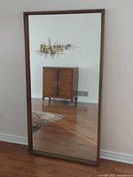Full view of the floor mirror showing upright placement against the wall reflecting a wooden cabinet and wall décor.