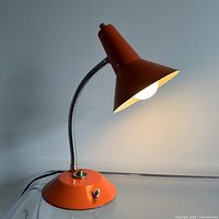 Photo showing the orange metal desk lamp turned on with light illuminating from the conical shade, highlighting the flexible metal gooseneck and circular base with switch and button.