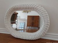 Front view of the white oval wicker mirror leaning against a wall, showing the mirror reflection of a room with furniture.