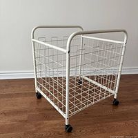 Front and side view of a vintage white metal wire rolling storage cart with caster wheels on a wood floor.