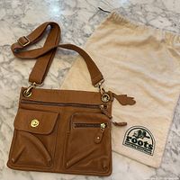 Butterscotch Roots crossbody bag lying flat on a marble surface with Roots dust bag next to it. Visible front zip pocket and flap pocket with gold twist lock.