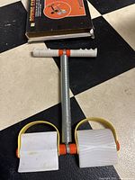 Rowing exerciser unit showing white foot pedals with yellow straps, metal shaft with spring, and white T-bar handle.