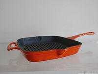 Side view of square cast iron grilling skillet with flame orange enamel exterior and black ridged interior cooking surface.