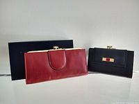 Two vintage ladies clutch wallets side by side, red and black, displayed in front of a navy Buxton box