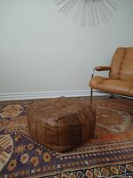 Square vintage leather patchwork ottoman set on a colorful patterned rug next to a modern leather chair with metal arms.