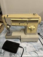 Front and side view of vintage cream-colored Singer Stylist sewing machine with foot pedal on marble surface.