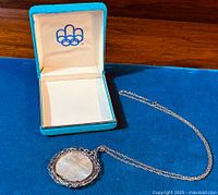 Photo showing the 1976 Canadian Olympic $10 silver coin pendant with chain and original turquoise-colored presentation box with Olympic rings inside the lid.