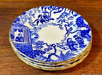 Stack of four blue and white Royal Crown Derby Mikado bread and butter plates with gold trim edges on wooden surface.