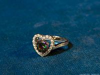 Ring angled to show heart-shaped mystic topaz center stone surrounded by white topaz stones on silver band against blue background.