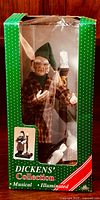 Front view of the boxed Dickens Collection Ebenezer Scrooge musical and illuminated Christmas decoration. The figure is visible inside green box with clear plastic window. Figure wears plaid robe and green nightcap, with candleholder beside it.
