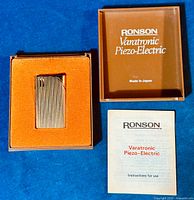 Vintage Ronson Veratronic lighter in its original box with instruction manual beside it, photographed on blue background.