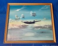Frontal photo of the framed signed oil painting showing WWII bomber flying above clouds with clear sky and light reflections on water below.