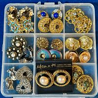 Clear overhead photo showing 16 pairs of goldtone vintage costume clip on earrings arranged in a plastic organizer box. Variety of designs including stones, pearls, and enamel. Monet brand visible on card for one pair.