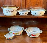 Photo showing the full set of six vintage Pyrex and Glasbake glass bowls with lids on a wooden shelf, including patterns and condition